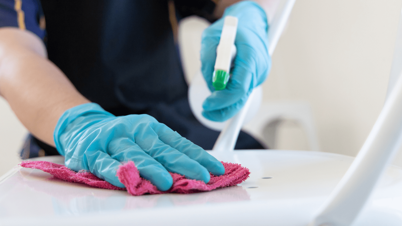 Person wearing blue gloves cleaning a white chair with a pink cloth and a spray bottle, illustrating residential cleaning services in Melbourne.