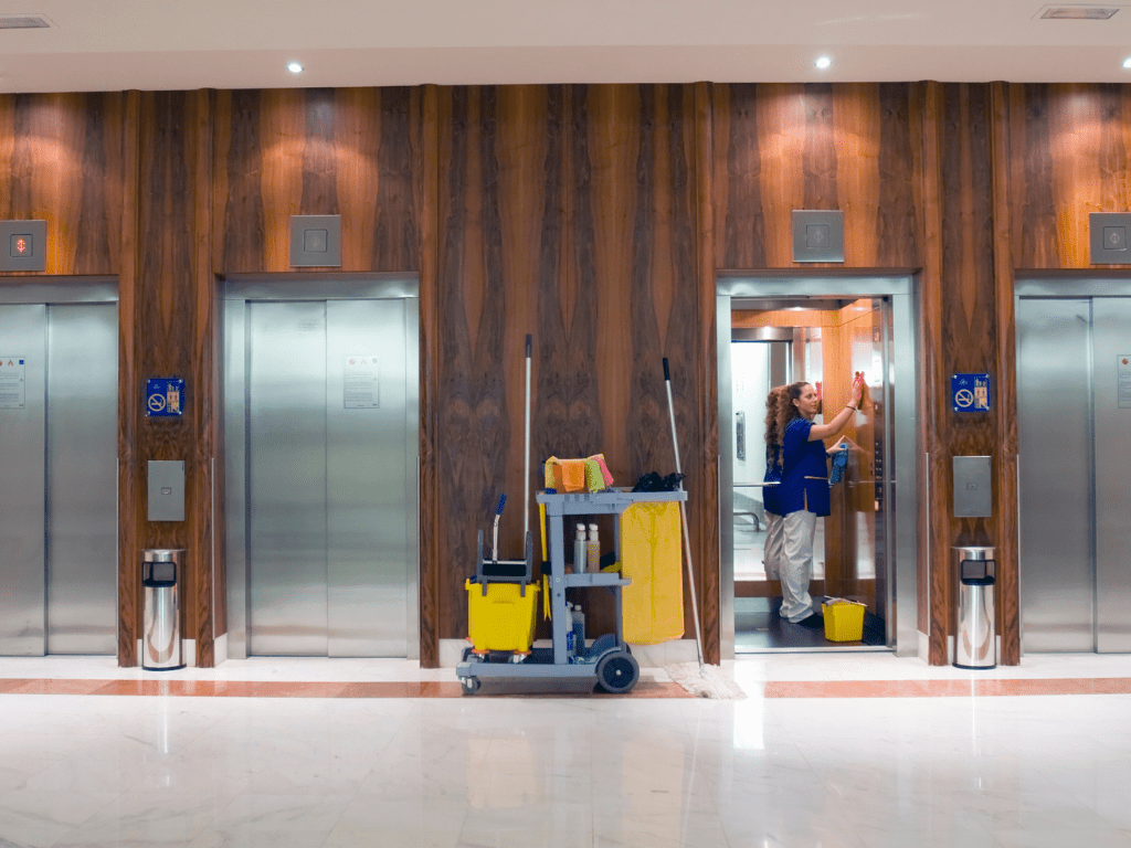 Strata cleaner in blue uniform sanitizing elevator doors in a modern building lobby with cleaning cart.